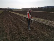 Andrea in red jacket sowing in a field with hilly landscape in the background.