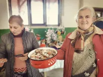 Two elderly women in a room. One woman is holding a bowl of sweet pastries, the other is watching. They are wearing warm clothes.