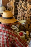 Bărbat cu pălărie de paie în fața unei grămezi de lemne și a unei mese cu roșii.
