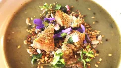 Egy tányér nagycsütörtöki leves ropogós darabokkal, magvakkal, fűszernövényekkel és lila virágokkal díszített krémes mártással.