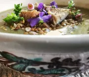 Soup plate with a creamy soup topped with crusty bread pieces, seeds and decorated with purple flowers and green herbs.