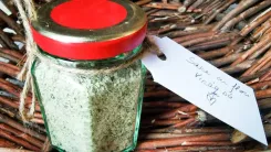 Glass jar with green content and red lid, on a wicker basket, with a pendant on which handwritten words.