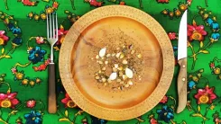 Ein Teller mit einer Portion Müsli, Nüssen und Samen in der Mitte, platziert auf einem bunten Tischtuch mit floralen Mustern. Ein Messer und eine Gabel liegen links und rechts neben dem Teller.