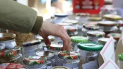 Andrea, reaching with her hand for a jar in her market stall full of various canned goods and jars.