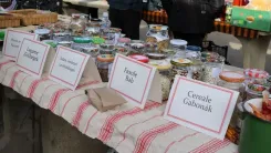 A market stall with a red and white striped tablecloth on which various canned goods and jars are displayed. In front of some products are signs with labels such as 'Tomato Basil' and 'Creole Goulash'.