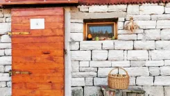 Eingang zum Saatguthaus mit einer holzvertäfelten Tür und einem Fenster, vor der weißen Ziegelmauer steht ein Korb mit Äpfeln auf einer Bank.