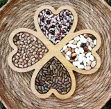A wooden cloverleaf design consisting of four heart-shaped compartments filled with various seeds and beans, placed in a round woven basket.