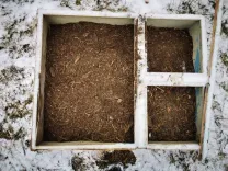 Drei verbundene quadratische Holzrahmen, gefüllt mit Humus, auf einem schneebedeckten Untergrund.
